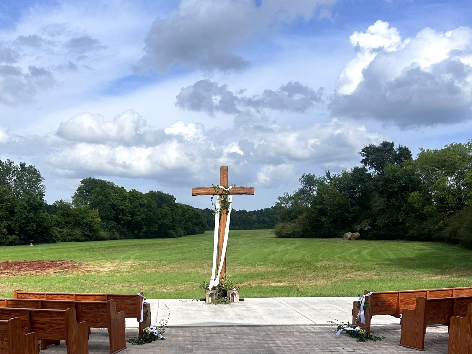 Wooden pews arranged on a paved court facing a handmade cross and an open green field